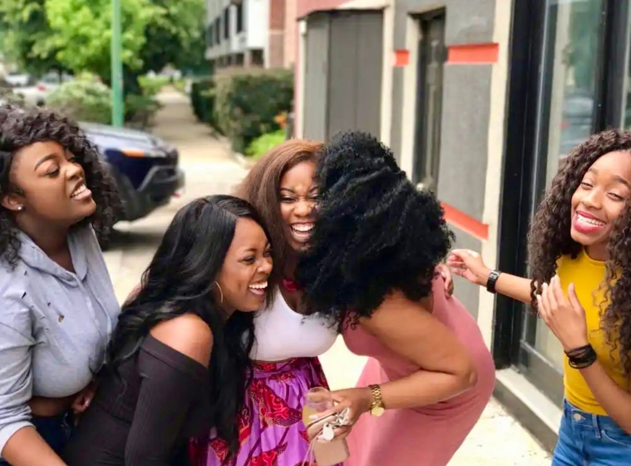 Five women laughing and embracing each other on a sidewalk near buildings and greenery.