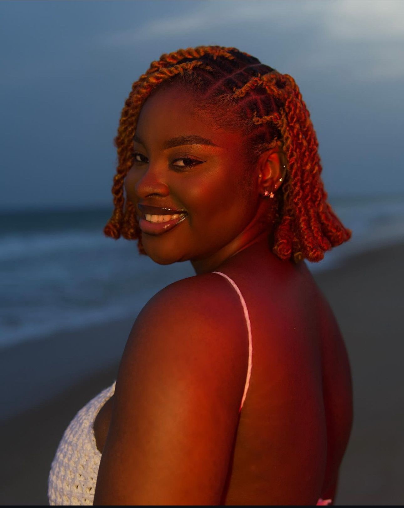 Person with twist hairstyle stands on the beach at sunset, wearing a white top.